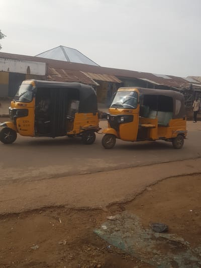 The image depicts two yellow and black "Keke Napep" auto-rickshaws parked on a paved road. The rickshaws are adorned with "ZRA" lettering. The background features a building with a metal roof and a sign for "ZYAB Carpentry & Services Nigl Ltd." The surrounding environment appears to be a dusty urban roadside in Tukur Tukur, Nigeria. There are no visible people in the immediate scene.