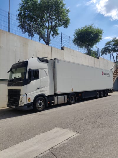 A white Volvo truck with a Lotos Baltica trailer is parked on an asphalt road in front of a concrete wall, with trees and a blue sky visible in the background. The truck has "GLOBETROTTER" written on the top of the cabin and a "VOLVO" logo on the front. The trailer has the "Lotos Baltica" logo and website printed on its side. The scene appears to be in an industrial or commercial area, with the concrete wall suggesting a boundary or retaining wall. The ground is an asphalt road with some cracks. The sky is partly cloudy. The location is identified as Mejorada del Campo, Spain.