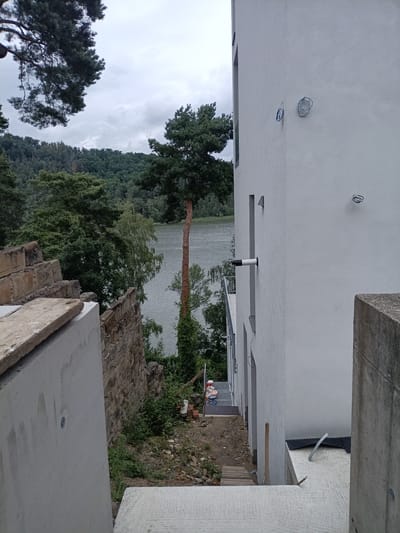 The image depicts an outdoor construction site at a building adjacent to a body of water, likely the Mácha Lake in Doksy, Czechia. The foreground shows unfinished concrete structures and some construction materials. A rough stone wall runs along the left side of the scene, separating the building from a steep, overgrown embankment leading down to the water.
A modern, multi-story building with a white facade is the primary structure visible on the right side of the image. Electrical conduits, some coiled, are visible on the exterior walls, suggesting ongoing electrical work. A metal railing and a drainpipe extend from the building towards the water. A set of metal stairs with a handrail is visible leading up towards the building from the lower ground.
On the stairs, a person wearing a construction helmet and orange vest is visible, seemingly overseeing the work or taking a break. Several paint buckets are placed near the base of the stairs. The surrounding environment is dominated by dense green trees, with a body of water reflecting the overcast sky in the background. The water appears calm, with a few ripples visible on its surface. The overall scene suggests a construction project nearing completion or in an advanced stage, set against a natural, lakeside landscape.