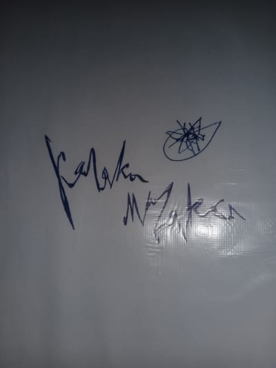 The image shows a white surface with blue ink markings. The markings include two sets of signatures and a doodle. The signature on the left appears to be "Karaka" or a similar phonetic interpretation, written in a cursive, stylized script. To the right of this signature is another signature, which is more difficult to decipher but appears to be "Mayaken" or a similar phonetic interpretation. Above and to the right of the signatures, there is a doodle resembling a starburst or a stylized flower within an oval shape. The white surface has a textured, possibly plastic or vinyl, pattern visible in places, suggesting it might be a whiteboard, a sign, or a covered surface. The lighting in the image creates shadows and highlights on the textured surface. The image does not contain any visual information that would allow for its location to be identified in the city of Gulbag, Uzbekistan, or any other specific geographical location.