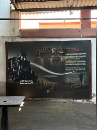 The image shows a large informational poster about the "Locomotora de Vapor 279" (Steam Locomotive 279), hanging on a concrete wall. The poster features photographs and text describing the history and significance of the locomotive, including its origin, function, and its role in Mexican history. A cropped view of the image shows the text and photos on the poster in detail. The text is in Spanish. The poster mentions that the locomotive is located in Cuautla, Mexico. There are no people visible.