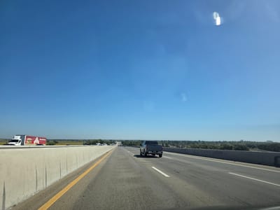 The image captures a view from a vehicle traveling on a highway under a clear blue sky. The road has multiple lanes, with a concrete barrier on the left and a low retaining wall on the right. A white semi-trailer truck with a red and white logo is visible on the left side of the road, and a gray pickup truck is in the right lane ahead. In the distance, trees and buildings can be seen, suggesting a developed area. The time of day appears to be late morning or early afternoon, indicated by the bright sunlight and long shadows. The weather is clear and sunny. The setting is a highway, likely in a rural or suburban area given the presence of trees and open fields beyond the roadside. There is no discernible emotion conveyed by the image, which presents a straightforward, objective view of the environment. The text on the side of the semi-trailer truck is visible, showing a logo with the word "OROWEAT" and an image of bread.