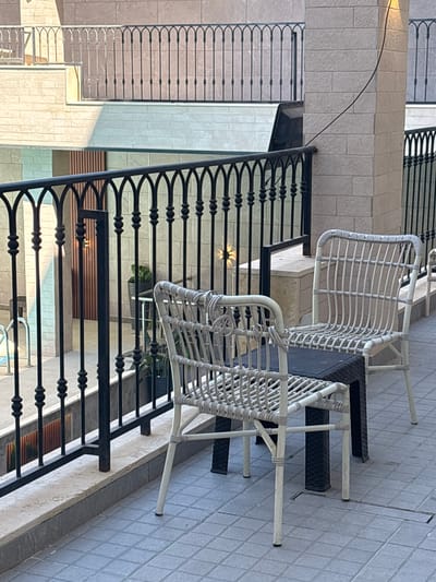 The image captures a balcony or patio area with a view of a building in Ayn al Basha, Jordan. Two white wicker chairs and a small black table are arranged on a tiled floor. A black metal railing with decorative arches runs along the front of the balcony, separating it from a lower level. The background features a stone-clad building with architectural details and some potted plants visible. Sunlight casts shadows on the scene, indicating it is daytime.