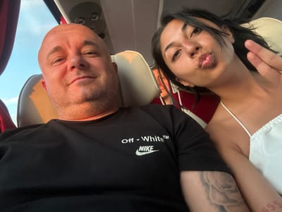A close-up selfie captures two individuals, a man and a woman, seated side-by-side in what appears to be the interior of a public transport vehicle, likely a bus, within the Municipal Unit of Lamia, Greece.
On the left, a bald man with a light stubble beard and fair skin looks directly at the camera with a faint smile. He wears a black short-sleeved t-shirt featuring the white text "Off - White™" above a white Nike swoosh logo. A large, light-toned tattoo, resembling a rose, is visible on his left bicep.
To his right, a woman with dark hair and medium skin tone leans her head towards the camera, pouting her lips in a "kissing" pose. She wears a white spaghetti-strap top. Her right hand is raised, displaying two fingers in a "peace sign" gesture, and a red "555" tattoo is visible on her right forearm.
The background shows beige upholstered seating, consistent with vehicle interiors. A window in the upper left reveals a clear blue sky, indicating daytime. Overhead vents and lighting fixtures are visible on the vehicle's ceiling, and a red curtain is partially seen on the far left.