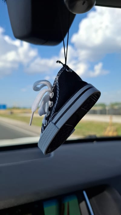 A black canvas shoe-shaped air freshener hangs from a black string, suspended from the car's rearview mirror mount. The shoe has white laces and white trim. The background is blurred, showing a bright blue sky with white clouds and a roadside landscape. The shoe is positioned in the foreground, and the car's dashboard is visible in the lower part of the image. There is no indication of the location being in Войводино, Bulgaria.