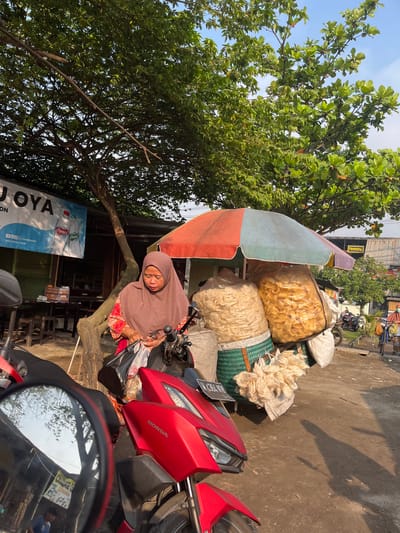 A middle-aged Indonesian woman wearing a brown hijab and a colorful patterned shirt stands next to a red Honda motorcycle. She is looking down and holding a black plastic bag filled with items. She appears to be at a roadside stall or market in Kedungkendo, Indonesia. The stall is adorned with a multicolored umbrella providing shade over large bags of what appear to be fried snacks or crackers. Behind the woman, a banner with text in Indonesian and English advertises "JOYA" and "cheers water". Trees with green foliage provide shade over the scene, and the ground is a dirt path. In the background, other market stalls and buildings are visible, along with a few other people and vehicles, suggesting a typical urban street scene. The lighting indicates it is daytime, with sunlight casting shadows on the ground.