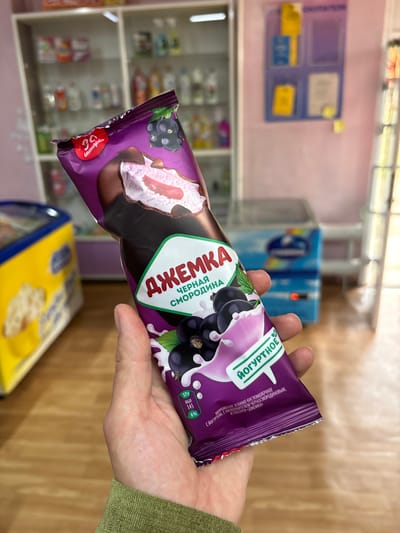 A person's hand holds a purple ice cream wrapper with a picture of blackcurrants and a depiction of the ice cream inside. The wrapper is labeled in Russian with the brand name "ДЖЕМКА" (Dzhemka) and "ЧЕРНАЯ СМОРОДИНА" (Blackcurrant). Below this, the words "ЙОГУРТНОЕ" (Yogurt) are visible. Nutritional information is present on the wrapper, showing "57г", "145 ккал", and "6%". The background suggests an indoor retail environment, possibly a small shop or convenience store, with shelves stocked with various products and refrigeration units. The flooring is wooden. The image is likely taken in the city of Nevon, Russia, given the Russian text on the product.