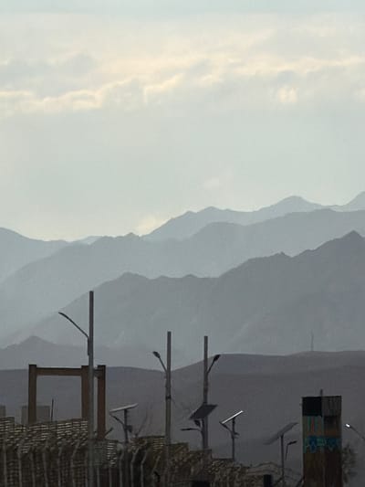 The image shows a hazy, mountainous landscape, likely in the city of Chumkar, India. In the foreground, there are several man-made structures, including a fence, a large rectangular frame, and streetlights. There are also solar panels visible. The mountains in the background are layered and faded due to the atmospheric haze. The sky is overcast with some lighter patches of clouds. There are no people visible in the image.