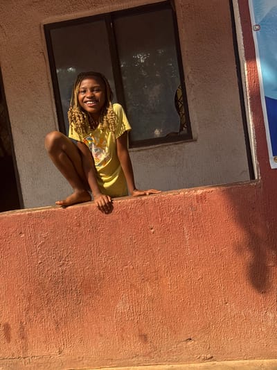 In Aguata, Nigeria, a young female is depicted in a squatting position atop a reddish-brown, textured wall. She is positioned towards the left side of the frame, facing the camera with a wide smile, exposing her teeth. Her hair is styled in numerous light-colored braids. She is barefoot and wears a yellow short-sleeved t-shirt featuring a colorful graphic design on the chest. Her hands are placed flat on the upper surface of the wall. Behind her, a building wall with a light-colored, textured finish contains a rectangular window with dark framing. The right pane of this window shows a visible crack or missing section. Reflections of foliage are discernible within the window panes. To the right of the window, a blue and white banner or poster is partially visible on a darker, reddish-brown section of the building, secured by a silver grommet at its upper right corner. The scene is illuminated by natural daylight.
