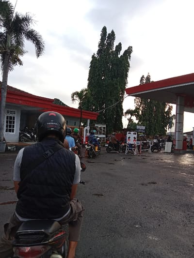 A person on a motorbike waits in line at a gas station. The person wears a helmet, vest, and grey shirt. Other motorbikes and people queue ahead. The gas station has a red roof and white pillars. A building with a red roof is on the left. Tall green trees and palm trees are visible in the background under a cloudy sky. The ground is asphalt and appears wet.