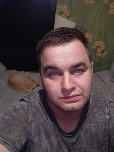 A selfie taken indoors by a man with short, dark hair and blue eyes. He is wearing a dark grey t-shirt. He has a light beard and stubble. To the left of the man, an orange cat is sleeping curled up on a bed. The background shows a black surface on the left and a green patterned wallpaper with yellow flowers on the right. The overall impression is of a casual, personal moment captured at home. The location is identified as Ketrysanivka, Ukraine.