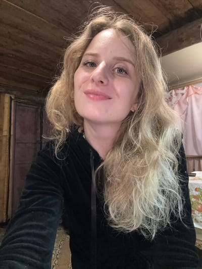 The image shows a close-up, selfie-style portrait of a young Caucasian woman with blonde, wavy hair, blue eyes, and light skin. She is wearing a black hooded jacket and is looking directly at the camera with a slight smile. The background appears to be an indoor setting, possibly a room with wooden paneling on the ceiling and a blurred pink curtain on the right side. There is also a glimpse of a table with a floral-patterned tablecloth. The lighting suggests an indoor environment, potentially at dusk or in a dimly lit room. Based on available information, the location is the city of Severnyy, Russia.