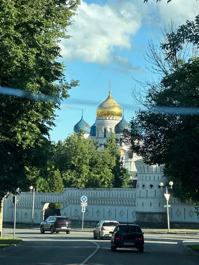 A sunny day in Dzerzhinsky, Russia, is depicted, with a view of the St. Nicholas Monastery. In the foreground, a black sedan and a white hatchback drive on a road. The black sedan is in the right lane, and the white hatchback is in the left lane, slightly ahead of it. The road is paved with asphalt and has white lane markings.
Behind the cars, a white stone wall with a patterned design runs along the street. Beyond the wall, the golden domes and blue spires of the St. Nicholas Monastery are visible, surrounded by lush green trees. The sky is blue with scattered white clouds. A traffic sign with a blue circle and a white cross is posted on the right side of the road, near the wall. Another traffic sign showing a white triangle with a red border is also visible on the right. The overall scene is one of a peaceful, sunny day in a city with historical and religious landmarks.