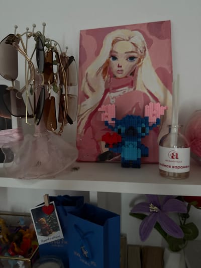 The image shows a shelf with various decorative items. On the left, a stand holds several pairs of sunglasses. Below the sunglasses, a pale pink tulle skirt is draped. To the right of the sunglasses is a painting of a blonde woman with blue eyes, rendered in shades of pink and yellow. In front of the painting, a blue pixelated figurine of Stitch from Lilo & Stitch is holding a red heart. Next to the Stitch figurine is a glass bottle with a white label that reads "AROMANTIQUE" and "солёная карамель" (salted caramel in Russian).
In the lower portion of the image, a small framed photo of a teddy bear in a Santa hat in front of a Christmas tree is clipped to a blue shopping bag. Another blue shopping bag is visible to the right. Further to the right, a purple flower with yellow stamens is placed in a white vase. The shelf appears to be located in a domestic setting, potentially a bedroom or living room. The specific location within Pod'yapolskoye, Russia is not discernible from the visual information provided.