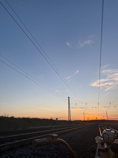 The image depicts a railway track at sunset in Schönebeck (Elbe), Germany. The sky is a gradient of blue and orange, with a few wispy clouds. Overhead, a network of power lines crisscrosses the sky, supported by tall metal poles. In the foreground, a section of the railway track is visible, covered in ballast. A yellow piece of machinery, possibly a rail tamper, is positioned on the right side of the track, partially in focus. The background shows a line of trees and distant structures, silhouetted against the setting sun. There are no people visible in the image.