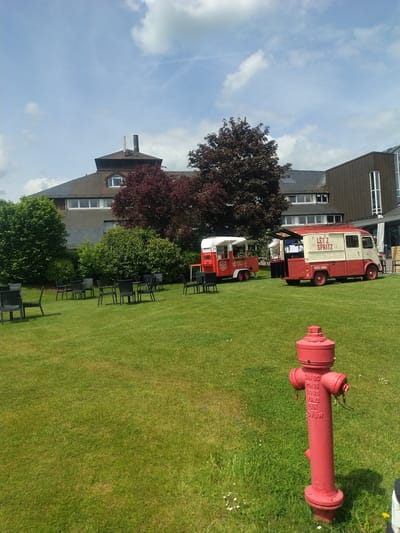 The image shows a grassy area near a building in Lenningen, Luxembourg. A red fire hydrant lies on the grass. In the background, two vintage vehicles converted into food trucks are visible. One has "LET'Z SPRITZ" written on its side, and the other has a logo that appears to be "BIKOLA." There are several chairs near the food trucks, seemingly set up for outdoor seating. The building is a multi-story structure with windows and a dark-colored exterior. Trees and bushes fill the space between the building and the food trucks. The sky is blue with some clouds.