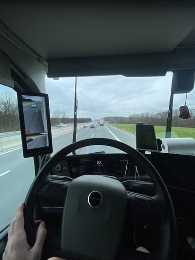 The image presents a first-person view from inside a Volvo heavy-duty truck's cabin, looking forward through the windshield onto a multi-lane highway near Mühlhausen, Germany. The sky overhead is overcast.
Inside the truck, the driver's left hand is visible on the lower left portion of the black steering wheel, which bears a silver "VOLVO" logo at its center. The digital dashboard display shows "82," indicating the vehicle's speed, likely in kilometers per hour. To the left of the windshield, a vertically mounted black digital side mirror monitor displays a real-time feed of the adjacent left lane, showing a white passenger car and the road surface. On the dashboard to the right, a navigation device is visible, displaying a map interface. A red and white air freshener, shaped like a Pepsi can, hangs from the overhead area on the right side. A roll of white paper towels rests on the far right side of the dashboard. Two vertical windshield wiper arms are positioned in the center of the windshield.
Outside the truck, the highway consists of at least two lanes for traffic moving in the same direction. Multiple vehicles, including other passenger cars and heavy-duty trucks, are visible ahead. The environment adjacent to the highway comprises green grassy verges, transitioning into dense forested areas further in the distance. Clear white lane markings delineate the asphalt road surface. The scene depicts an active driving situation on an open road.