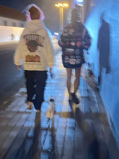 This blurred, low-light image captures two individuals and a small animal walking away from the viewer on a paved surface in San Fernando, Spain. The subject on the left wears a white hoodie with a rear graphic depicting text (possibly "MONSTER") and illustrations resembling pumpkins or squashes, dark trousers, and light-colored shoes; their head shows a diffuse, light, possibly pinkish form. The subject on the right wears a dark, patterned jacket or dress extending to mid-thigh, exposing legs, and dark footwear; their hair is gathered. A small, light-colored animal, resembling a cat, walks ahead of the second subject on the block-paved ground. A wall to the right is illuminated with blue light, casting distinct, elongated shadows. In the background, a multi-headed streetlamp emits yellow light, and a building facade is partially visible on the left.