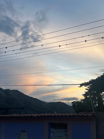 The image captures a serene sunset scene in Villa de Cura, Venezuela. The sky is painted with soft hues of blue, gray, and a gentle orange glow, indicating the approaching twilight. Several parallel power lines stretch across the frame, adorned with small, spiky objects, possibly nests or seed pods.

In the middle ground, a dark, silhouetted mountain range dominates the left side of the image, with subtle contours and scattered greenery visible on its slopes. A hint of a distant transmission tower can be observed on the far right of the mountain range. Below the mountains, a red-tiled roofline runs horizontally across the lower portion of the image, partially obscuring a blue wall with white architectural details. A lush, dark green tree canopy extends into the frame from the right, its branches reaching upwards. The overall atmosphere is peaceful and tranquil, characteristic of a rural or semi-rural setting at dusk.