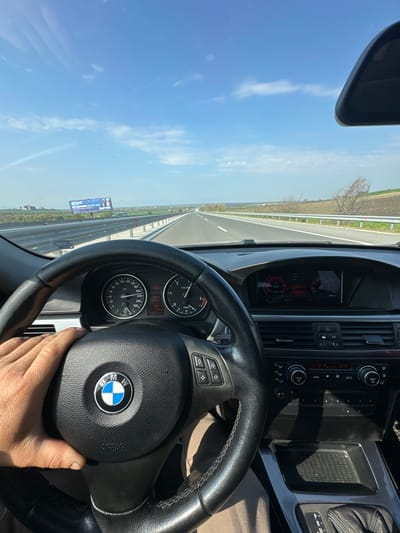 The image depicts the interior of a BMW vehicle, specifically the driver's perspective. The steering wheel, with the BMW logo prominently displayed, is the central object. The driver's hand is visible holding the steering wheel. The dashboard, including the speedometer and other gauges, is in focus. The car is traveling on a highway, as evidenced by the road visible through the windshield. A roadside billboard displaying the text "NETTHALAD" is on the right side. The sky is clear and blue. Based on the identified text on the billboard, the location is Rupkite, Bulgaria. The car is showing a mileage of 213.9 kilometers. The temperature is showing 21.5 degrees Celsius. The speedometer indicates a speed of 209 kilometers per hour.