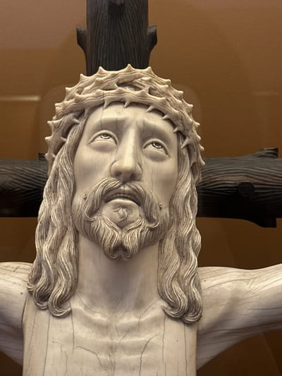 A close-up, horizontally oriented photograph captures a meticulously carved sculpture of Jesus Christ on the cross, originating from Tepotzotlán, Mexico. The sculpture is crafted from a pale, smooth material, likely ivory or aged bone, displaying fine lines and subtle cracks consistent with its age. Jesus's face is depicted with profound solemnity, featuring open eyes gazing slightly upwards, a finely sculpted beard, and long, flowing wavy hair intricately rendered. A prominent crown of sharp thorns encircles his head, with each thorn distinctly carved.
The cross to which the figure is affixed is made of a dark, textured wood, exhibiting distinct vertical grain patterns. The sculpture is presented against a plain, warm brown background, suggesting an indoor setting such as a museum, church, or gallery. The lighting subtly illuminates the sculpture, emphasizing its intricate details and contours while casting soft shadows that add depth to the figure. No activity is taking place, as the image presents a static, reverent representation of the crucifixion. There is no visible text in the image.