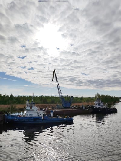 The scene depicts a waterway in Ozernoe Ust'e, Russia, with two tugboats and a barge equipped with a crane. The tugboats are positioned on either side of the barge, appearing to be moored. The crane is centrally located on the barge, with its boom extending upwards. The waterway's surface reflects the sky and vessels. The shoreline is visible in the background, lined with trees and vegetation. The sky is overcast with clouds, obscuring the sun.