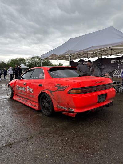 The image shows a red sports car with the logos "CERAMIC PRO" and "BN Sports", parked on a wet surface. The car appears dirty, suggesting it may have recently been used in a race. The car is parked beneath a tent with the word "INTELEGENT" printed on the side. Several people are standing around in the background. The license plate says “INTELEGENTOFF” and the user’s Instagram handle is printed on the window. The image was likely taken at a car event in Pavlovskaya, Russia, based on the Cyrillic text "ДЕТЕЙПИНГ ЦЕНТР" (Detailing Center) on the car.