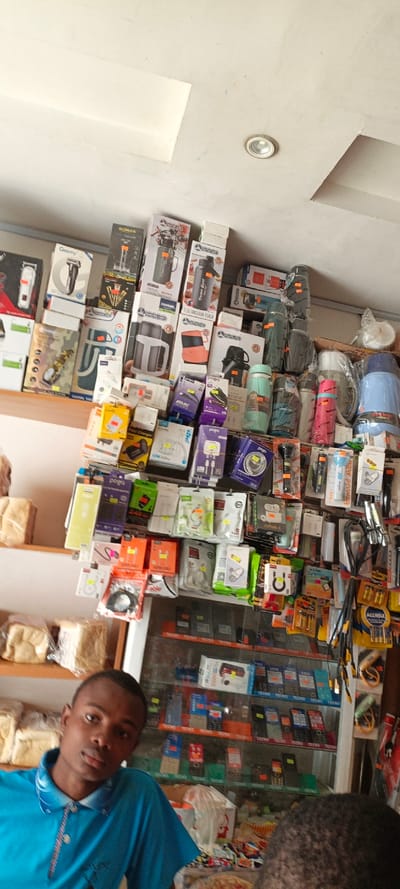 A brightly lit shop displays a variety of items. The upper shelf holds boxes of water flasks and grooming devices. Below, packages of phone chargers, cables, and batteries are arranged. A glass cabinet features numerous Tecno-branded feature phones. Two men are visible in the foreground, one wearing a blue polo shirt looking towards the camera.