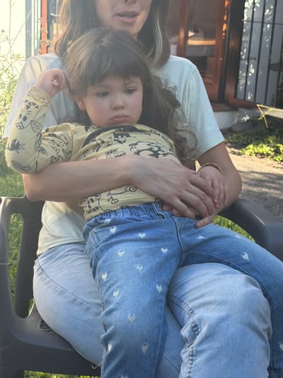 A young child rests on an adult's lap. The child is wearing a patterned yellow shirt, while the adult is wearing light-colored clothing and jeans. The adult's jeans have small heart patterns. They are sitting in a gray plastic chair outdoors in the city of Tolosa, Argentina.