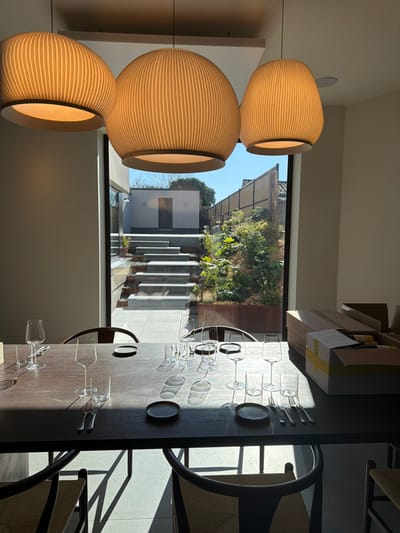 The image displays an indoor dining area with a panoramic view of an outdoor terraced garden, situated in Marche-lez-Écaussinnes, Belgium. Three prominent, unlit, ribbed, light-colored pendant light fixtures hang from the ceiling above a long, dark wooden dining table. The table is partially set with multiple empty stemmed wine glasses, stemless water glasses, small dark circular plates, and silverware (forks and knives). Strong natural sunlight streams through a large window, casting sharp shadows of the table settings and chair frames across the table surface and floor. On the right side of the table, two open cardboard boxes containing unidentified contents are present.

The outdoor scene, visible through the window, features a multi-level terraced garden. At the rear, a light-colored outbuilding with a dark door and two wall-mounted sconce lights is visible. Grey rectangular stone steps descend across several paved levels. Lush green foliage grows in dark, rectangular planters positioned along the right side of the garden, which is bordered by a tall, slatted wooden fence. The sky above is clear and blue. No individuals are present or engaged in any activity within the observed scene.