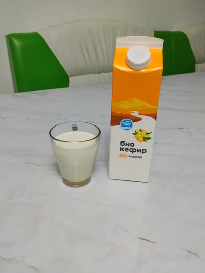 The image displays a glass of kefir and a carton of the same product on a marble-patterned table. The kefir carton is primarily orange and white with a depiction of a landscape featuring mountains and a river. Visible text on the carton includes "белая река," "био кефир," and "+ бифидобактерии." A small floral graphic, possibly representing vanilla, is also present. To the left of the table, a portion of a sofa or bench with green and white upholstery is visible. The scene appears to be indoors, with neutral lighting, suggesting it could be daytime. The arrangement of the objects suggests a simple still life presentation. The overall impression is one of everyday products displayed in a domestic setting. No people are visible in the image. The location context of Almaluu, Kyrgyzstan, is provided but not visually evident within the frame.