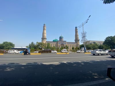 The image shows a wide, paved road in front of a large, ornate mosque with two tall minarets and a green dome. Several vehicles are visible on the road, including a blue and white bus, a white car, a black SUV, and a blue auto-rickshaw with a person riding it. The mosque is decorated with intricate blue and white tilework. Trees line the area between the road and the mosque. The sky is clear and blue. This scene is located in Andijan City, Uzbekistan.