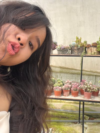 A close-up, selfie-style portrait shows a young person with dark, flowing hair puckering their lips. The person has light brown skin, dark eyes, and full lips. The background, slightly blurred, reveals a collection of potted plants on tiered shelves, including cacti and succulents. The scene likely takes place in a greenhouse or outdoor garden setting. The specific location of Măng Đen, Vietnam, is not evident in the image itself.