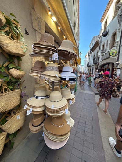 This is a vertical, eye-level shot of a street market stall in Tossa de Mar, Spain. The scene is brightly lit, suggesting it is daytime, likely sunny. The stall displays a wide variety of straw hats and woven bags. The hats are stacked and hung, showcasing different styles, colors, and embellishments, including some with decorative bands and shells. The bags are also woven, with some featuring handles. In the background, a cobblestone street is visible with several people walking and browsing. The architecture of the buildings lining the street appears to be traditional Mediterranean style. Some signs are visible on the buildings, including one that reads "GUINNESS PUB" and another with a cartoon chicken that says "CHOUFFE". The overall atmosphere seems relaxed and summery, typical of a tourist town. The focus is on the array of hats and bags, with people in the background serving to provide context for the setting.