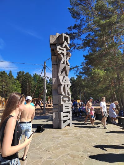 This image depicts a public space in Solnechnoye, Russia, on a clear, sunny day. A large, grey stone monument with Cyrillic letters spelling "СОЛНЕЧНОЕ" (Solnechnoye) stands prominently in the foreground. The monument is composed of stacked, irregularly shaped stone blocks, each carved with a letter. A paved pathway made of irregular stone slabs surrounds the monument.
Several people are visible in the scene. In the foreground left, a woman with long brown hair is looking at her phone. To her right, a man with a white cap and a bare torso, wearing denim shorts, stands facing away from the camera. Behind them, other individuals and groups are walking along the pathway and seated on benches. A playground structure is visible in the background, suggesting a family-friendly recreational area. Tall pine trees line the perimeter of the area, casting shadows on the ground. A utility pole with electrical wires and security cameras is situated near the monument. The overall impression is of a well-attended, outdoor public gathering space.