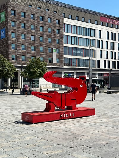 This media file shows a large, bright red metal sculpture in the shape of a stylized alligator, placed on a paved plaza. The sculpture is positioned diagonally across the frame, with its head and jaws in the foreground. Text on the sculpture's base reads "NÎMES".

In the background, a tall, modern building complex is visible. One section of the building has a facade of horizontal wooden slats, with many windows. Another section of the building is lighter in color and features a repeating pattern of windows. Signs for "Midi Libre" and "MAISON de MA rég" are visible on this section. Further in the background, another part of the building is labeled "HOTEL IBIS". Below the wooden-slatted facade, there are banners for "ibis STYLES" and "ibis budget".

Two people are visible in the scene. One person is standing near the red sculpture, and another is seated on the ground further behind the sculpture. The scene appears to be outdoors on a sunny day, indicated by the clear blue sky and the strong shadows cast by the sculpture and the building. The overall impression is of an urban public space with a modern artistic element.