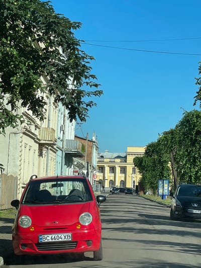This image depicts a street scene in Stryi, Ukraine, under a clear blue sky, suggesting it is daytime and fair weather. The perspective is from inside a vehicle, looking forward and slightly to the right. In the foreground, the bright red hood and windshield of a small car are visible. The car has a license plate that reads "BC 6604 XB" and appears to be parked or moving slowly.
To the right of the red car, a road is lined with shadows cast by trees and buildings, indicating a sunny day. Several other cars are visible further down the street, appearing to be in traffic. The buildings lining the street are multi-story structures with architectural details, some appearing older and ornate, while others are more modern. One prominent building in the middle ground is painted a pale yellow.
On the left side of the image, lush green trees with dense foliage partially obscure the view of the buildings and the sky. The branches of these trees hang over the street. In the background, a tall dark-colored car is parked or driving on the road, its license plate is visible but illegible. Further down the street, a blue sign with some text and symbols is visible. The overall impression is a typical urban street scene during the day, with cars, buildings, and trees contributing to the environment.