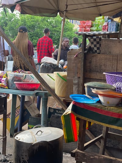 This image captures a vibrant and busy outdoor market scene, likely taken during the daytime. The perspective is from within or very close to a market stall, looking outwards towards people and other stalls. The scene is characterized by a mix of makeshift wooden structures, goods for sale, and several individuals.
In the foreground on the left, a large, light-colored umbrella or canopy provides shade. Below it, in the lower left, a person is visible from the back, wearing a black cap and sporting long, light-colored braids. Next to them, further into the mid-ground, another person is seen from the back, wearing a red and black plaid shirt.
Central to the mid-ground, a man with dark skin is seated on what appears to be a motorbike, or at least has a motorbike in front of him. He is wearing a dark-colored shirt and has an animated expression, with his mouth open, looking towards the right side of the frame. Behind him, another person in a lighter-colored shirt is partially visible. In the background, beyond the people, there are green trees and a building with the word "UNIVERSITY" partially visible on its facade.
The right side of the image is dominated by a market stall setup. It consists of rough wooden shelves and supports packed with various items. Towards the top right, a purple plastic basket, along with white, orange, and blue plastic bowls or basins, are hanging or displayed. Below these, a colorful fabric with distinct red, green, yellow, and black stripes is draped over a wooden beam.
On the shelves and surfaces of the stall, numerous items are stacked and displayed. These include brightly colored packets of goods, possibly snacks or small household items, in green, pink, and blue clear plastic packaging. A black and white checkered cloth is also visible on one of the wooden surfaces. Lower down in the stall area, there are yellow and clear plastic buckets, one with a green handle, and a red plastic colander or basket. Large metal pots with lids are prominently placed in the lower right corner, suggesting cooking or serving food. Several cardboard boxes are stacked and scattered around the stall, with visible text including "BUTTERS," "Galls," "PACK," and smaller text that seems to include "Rite."
The overall impression is one of a lively, functional marketplace with goods readily available and people moving through or engaging with the vendors. The lighting suggests a bright day, possibly with some cloud cover or shade from the canopies.