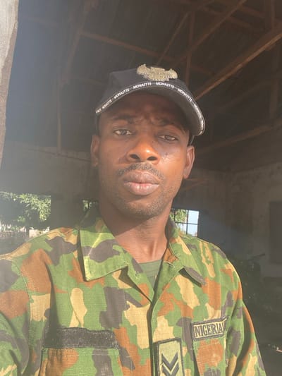 The image displays a close-up, selfie-style photograph of a dark-skinned man. He is looking directly at the camera with a neutral expression. Strong, natural sunlight illuminates the right side of his face and shoulder, creating bright highlights and shadows.
The man is wearing a dark blue or black cap with white text repeated around the brim, which clearly reads "MONAITTE". There is also a metallic, gold-colored emblem on the front of the cap, possibly depicting an eagle or a similar crest. He has short, dark facial hair, including a beard and mustache.
He is dressed in a military camouflage uniform, characterized by a pattern of green, brown, and black shapes. On his right shoulder (viewer's left side), a rectangular patch is visible with the word "NIGERIA" written in white capital letters on a dark background. Below this, there is another patch displaying a rank insignia, which consists of two gold or yellow chevron-like symbols pointing to the right. A dark, rectangular velcro patch is also visible further down on the uniform sleeve.
The background behind the man appears to be an interior space with a rustic or unfinished look. Wooden beams or rafters are visible overhead. Bright daylight is streaming in from an opening in the background, possibly a window or a gap in a wall, and some green foliage can be faintly seen outside. The strong illumination suggests it is daytime and sunny.