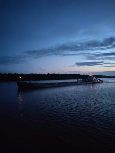 A barge, illuminated by its deck lights, travels on a dark body of water, likely a river or canal. The sky above is a deep blue, transitioning to lighter hues near the horizon where scattered clouds are visible. A dark line of trees forms the distant shoreline. The overall scene suggests dusk or early dawn in a rural or natural setting.