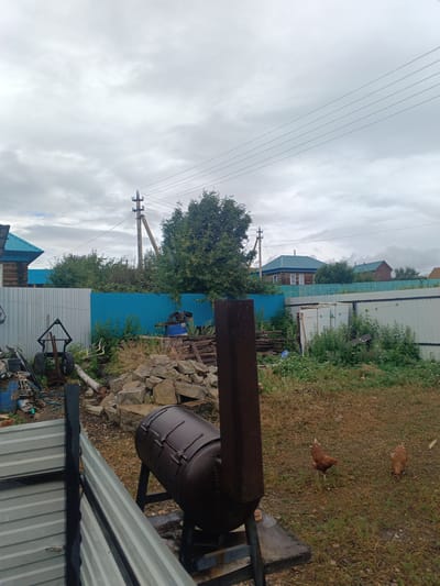 The image depicts a backyard scene in Iltebanovo, Russia. In the foreground, a large, dark brown metal structure resembling a wood-fired oven or boiler is positioned on a concrete base. A dark metal fence is visible to the left, and a pile of large rocks is located behind the oven. Further back, a bright blue fence separates the yard from the neighboring property, beyond which a wooden house with a blue roof and a modern metal structure can be seen. Trees and foliage surround the houses, and power lines are strung across the overcast sky. Two brown chickens are foraging on the grassy ground in the right portion of the image. The environment appears to be rural or semi-rural, with evidence of construction materials and various outdoor equipment.