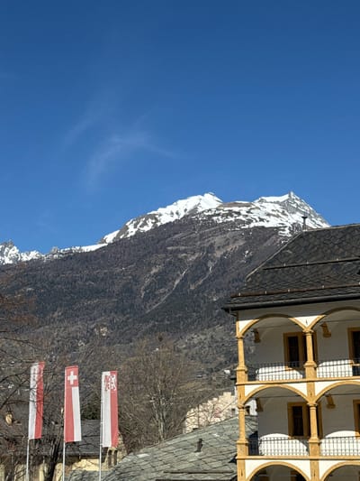 The image displays an outdoor scene in Visp, Switzerland, under a clear blue sky with minimal, wispy clouds. Prominently featured in the background are snow-capped mountains, their lower and mid-slopes densely covered with dark, bare trees, suggesting a winter or early spring season.

In the right foreground, a multi-story building, identified as a section of the Stockalper Palace, presents a white facade accented by yellow-gold columns and trim, forming a series of arched loggias on at least three visible levels. Dark, rectangular windows are set within these arches, and black wrought-iron railings adorn the balconies. The building is topped with a dark, possibly slate, pitched roof, from which a metal chimney pipe protrudes.

To the left of the frame, three vertical flags are displayed on poles. From left to right, they are: a white flag with a partial red border, the national flag of Switzerland (red field with a white cross), and the flag of the Canton of Valais (red and white vertical stripes with a partial emblem in the upper hoist). Portions of other buildings with dark pitched roofs are visible in the mid-ground, situated amongst bare deciduous trees. No people or active interactions are depicted in the scene.
