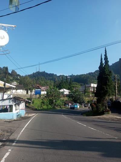 The image presents a street scene in a rural area, potentially located near a mountainous region. The road is paved and curves slightly to the right. A small building, possibly a shop or stall, is situated on the left side of the road. There are trees and vegetation present, along with buildings in the background that suggest a small community or village nestled into the landscape. Telephone poles and power lines are visible overhead. The sky is clear blue, indicating fair weather. A sign with Arabic script is present on the left. The presence of vehicles indicates active road traffic. The mountain in the background has cell towers on top. The image does not contain enough information to confirm or deny the location as Phouviang-Gnai, Laos.