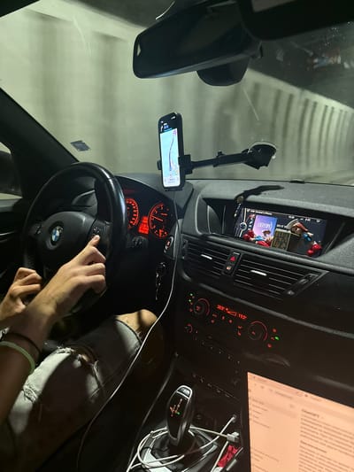 The image shows the interior of a car, likely a BMW, at night. The car appears to be in motion, as indicated by the blurred background of a tunnel. The driver's hands are on the steering wheel, and a smartphone is mounted on the windshield displaying navigation. The car's infotainment system is visible, showing music playback and a phone connection. A laptop is open on the passenger seat, displaying a webpage. The scene is consistent with a car traveling through a tunnel on a road near Dolna Vasilitsa, Bulgaria.