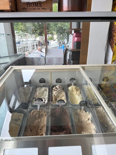This is a vertical, eye-level medium shot of an open ice cream freezer display in a shop. The freezer is filled with various tubs of ice cream. The flavors visible include Vanilla Madagascar, Heartma, Speculoos, Chocho Berries, Caramel, Stracciatella, Tiramisu, and others that are not fully legible. The brand name "κρι κρι" is visible on the labels of many of the tubs.
To the left of the freezer, a wooden crate with the text "HOME There's no place like Home" is visible. Behind the crate and partially visible through the glass of the freezer, the exterior scene is blurry, showing greenery, trees, and structures. There are stacks of white boxes, possibly for deliveries or storage, and what appear to be white chairs and tables, suggesting an outdoor seating area or a patio. The overall impression is a bright, daytime scene, likely in a warm climate, given the ice cream display and outdoor setting. The activity taking place is the display and sale of ice cream. There are no people clearly visible in the foreground, but a blurry figure might be present in the background, near the stacks of boxes.