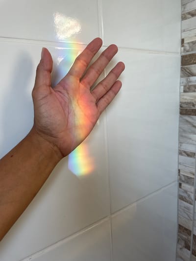 A person's left hand is positioned against a white tiled surface, likely a wall. A band of rainbow-colored light falls across the palm and fingers, originating from the upper right and extending towards the left. The arm extends downward and out of frame towards the lower right. A decorative tile border is visible at the top. The location is not specified.