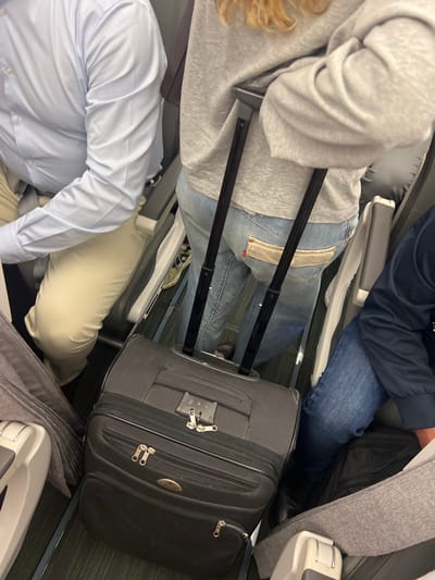 The image captures a close-up view of passengers seated in an airplane cabin, with their luggage positioned between seats. On the left, a person wearing a light blue button-down shirt is partially visible, their arm resting on their lap. Next to them, a person in a grey hoodie and blue jeans is seen from the waist down, holding onto the handle of a black rolling suitcase. This suitcase is directly in front of them, taking up a significant portion of the frame. To the right, another passenger's legs, clad in blue jeans, are visible, along with a dark jacket. The floor of the cabin appears to be covered in a dark carpet with faint green stripes. The overall scene suggests a typical situation during air travel, with passengers and their belongings arranged in a confined space. The location of Moreira, Portugal, is not discernible from the visual information provided in the image.