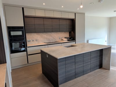 The image shows a modern, empty kitchen with a large island. The kitchen features sleek cabinetry in a neutral color, with dark wood accents on the island and some of the wall cabinets. Integrated appliances, including double ovens, are visible. The countertops appear to be made of a light-colored, veined stone. The island has a sink and is topped with the same stone as the countertops. The flooring is a light-colored wood-effect tile. The lighting appears to be a combination of recessed ceiling lights and under-cabinet lighting, suggesting the photo was taken during the day or evening with artificial lighting. There are no people visible in the image. The scene depicts a clean, contemporary kitchen, likely in a residential setting in Waterford, Ireland. The overall impression is one of spaciousness and modern design.
