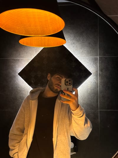 The image depicts a man taking a selfie in a bathroom. He is wearing a grey hoodie over a black shirt. The bathroom has black tiled walls and floor. There are two large, round, orange light fixtures on the wall. The man's reflection is visible in a diamond-shaped mirror.
While the image is stylish, there is no way to determine if this image was taken in Tsakhkadzor, Armenia. There are no details to connect the image to this city.