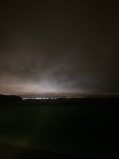 A night-time panoramic image depicts the landscape of Saint-Nom-la-Bretèche, France, viewed from a distance. The upper two-thirds of the frame are dominated by a heavily clouded, dark gray sky. Significant light pollution emanating from the distant settlement illuminates the underside of the clouds, creating a diffused, yellowish-white and orange glow along the horizon line. A continuous line of small, blurred artificial lights, consistent with an urban or suburban area, extends horizontally across the middle distance, indicating the location of Saint-Nom-la-Bretèche. The foreground and midground, comprising the lower third of the image, are rendered in deep shadow, showing indistinct dark land, possibly open fields. A dark, silhouetted landmass or treeline is visible on the far left, bordering the horizon. No specific individuals, identifiable objects, or discernible actions are present in the immediate vicinity or detailed within the distant town.